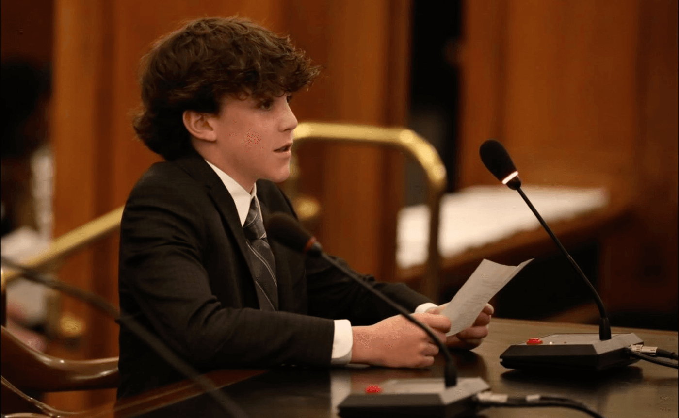 A young man with curly brown hair, wearing a dark suit and tie, sits at a desk with microphones, reading from a piece of paper in a wood-paneled room.