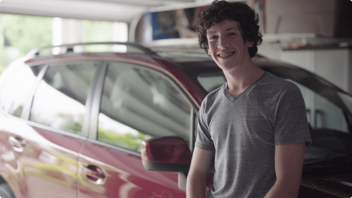 A young man with curly hair and braces smiles while posing in front of a red car parked in a garage.