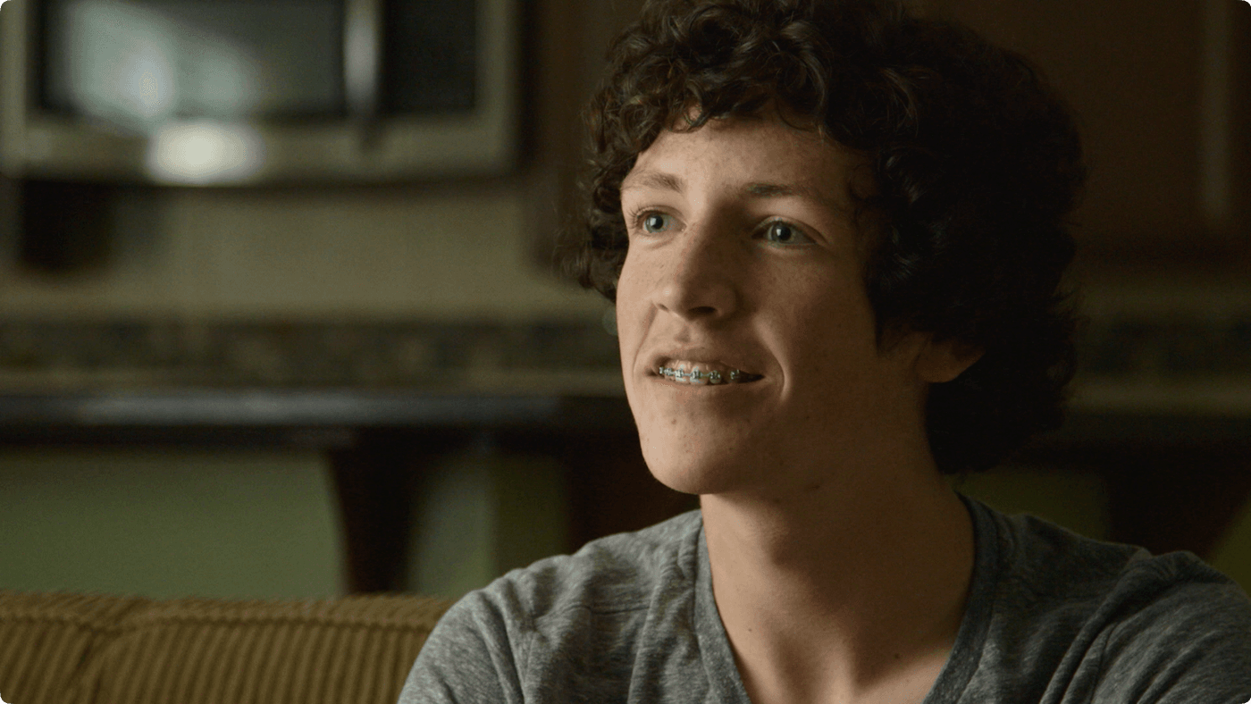 A close-up shot of a young man with curly brown hair and green braces looking slightly off-camera while wearing a grey t-shirt.