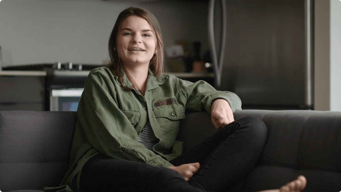 A young woman with long hair and a smile sits on a dark gray couch, wearing a green button-down jacket with a U.S. Army patch over black jeans.