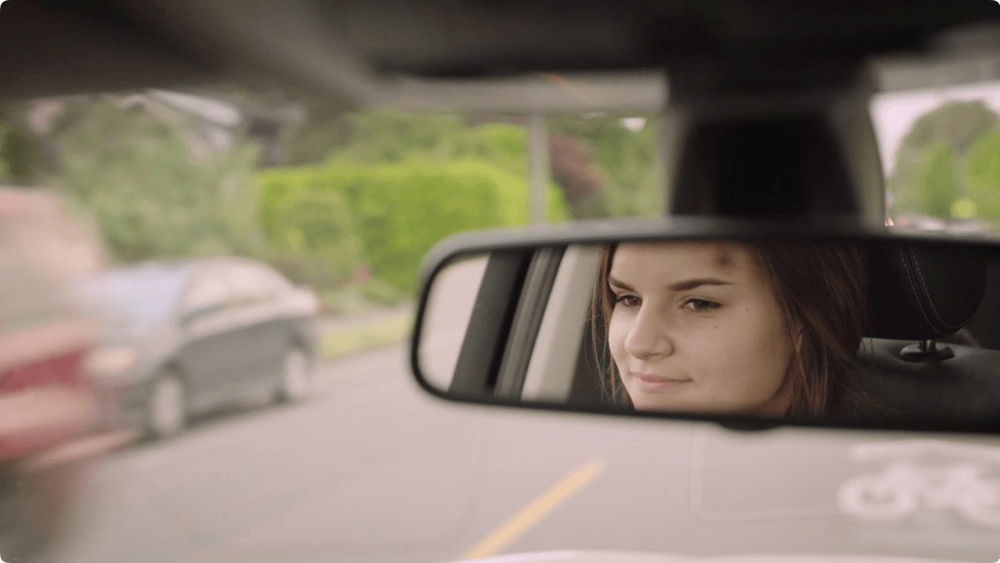 A young woman with a bruise on her forehead looks into the rearview mirror of a car while driving on a suburban road.