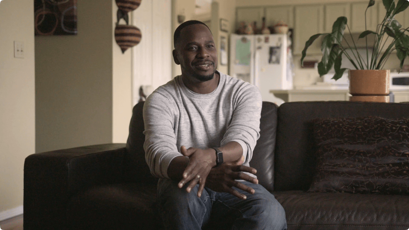 A man in a light grey sweater and blue jeans sits on a dark leather couch in a living room with his hands folded in his lap.
