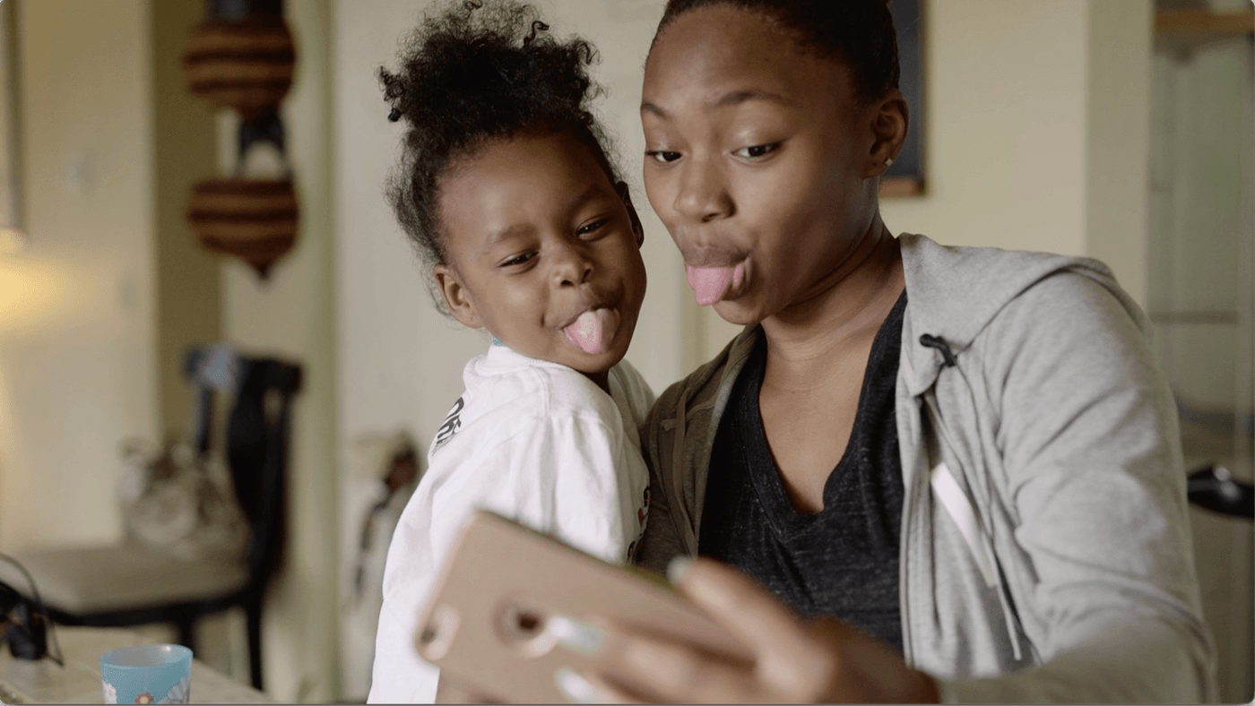 A woman and a young girl make silly faces, both sticking out their tongues, while taking a selfie with a smartphone held by the woman.
