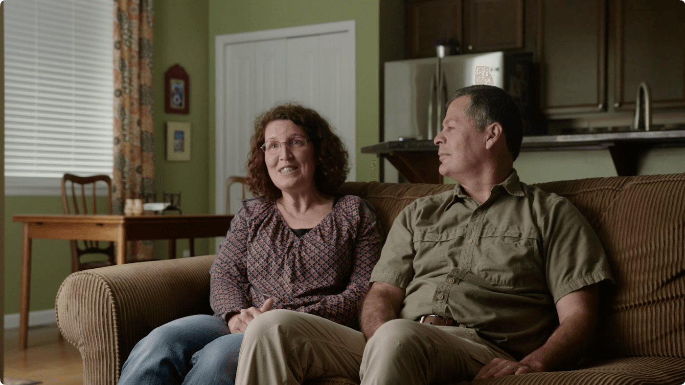 A middle-aged woman with curly reddish-brown hair and glasses talks while sitting next to her husband on a brown corduroy sofa in a home with green walls.