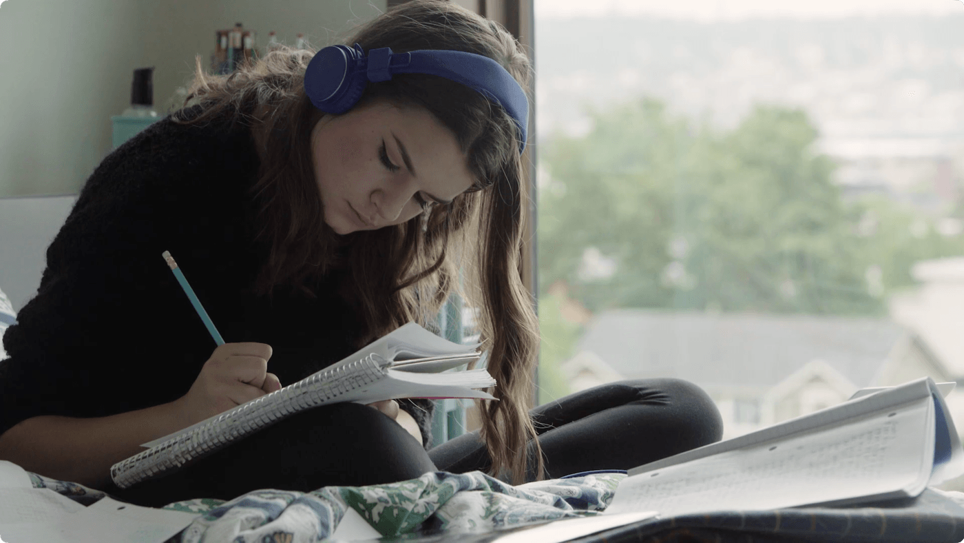 A young woman with long hair and blue headphones writes in a spiral notebook while sitting on a bed next to a window overlooking a neighborhood.