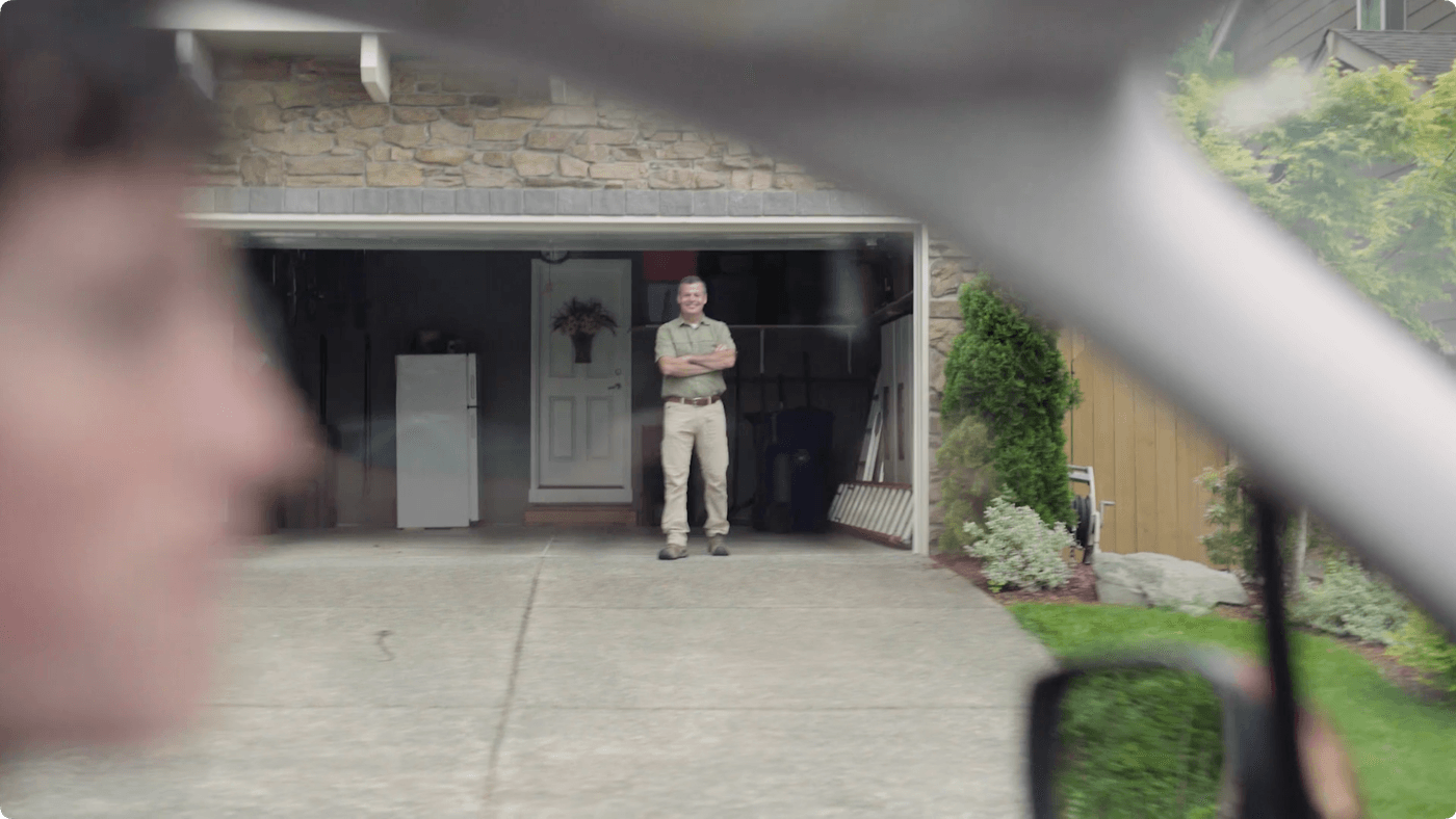 A man with his arms crossed stands in the center of an open garage, viewed from inside a car with a blurry person in the foreground.