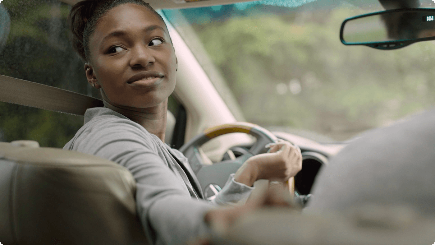 A young Black woman with her hair in an updo sits in the driver's seat of a car, smiling and looking back toward the passenger seat while holding out her hand.