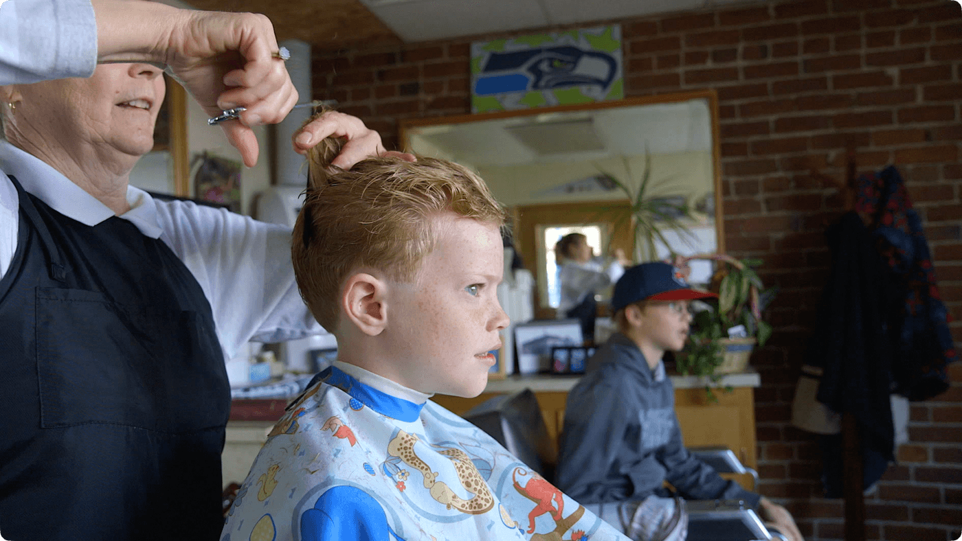 A hairdresser trims the blonde, freckled hair of a young boy wearing a patterned cape while another boy waits in a chair in the background of a brick-walled barbershop.