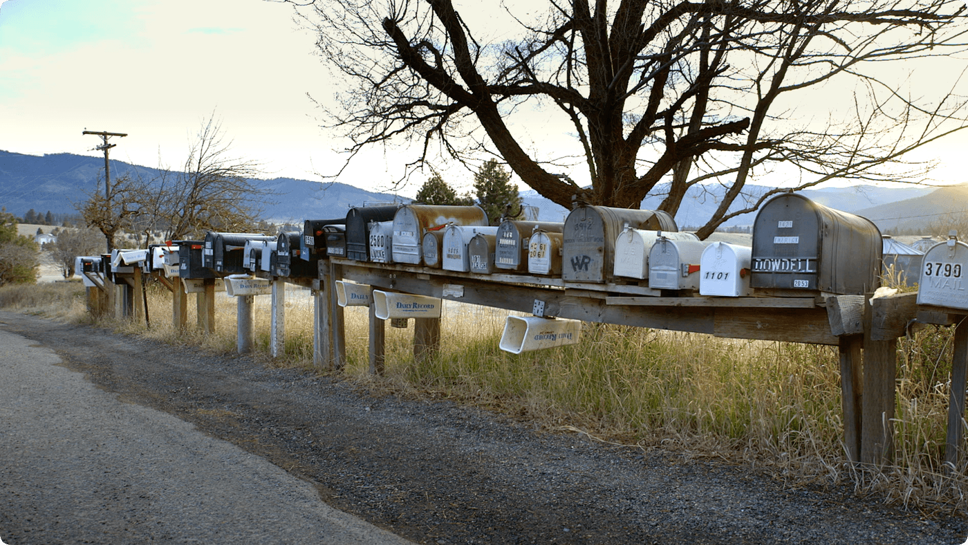 A long wooden rack holds a diverse row of assorted residential mailboxes and newspaper tubes along a gravel road, with a large leafless tree and distant mountains in the background.