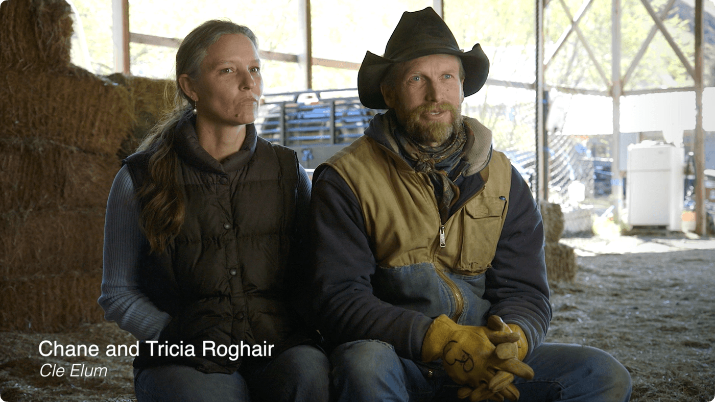 A man wearing a cowboy hat and work gloves and a woman with her hair pulled back sit together in a barn, with text at the bottom identifying them as Chane and Tricia Roghair from Cle Elum.