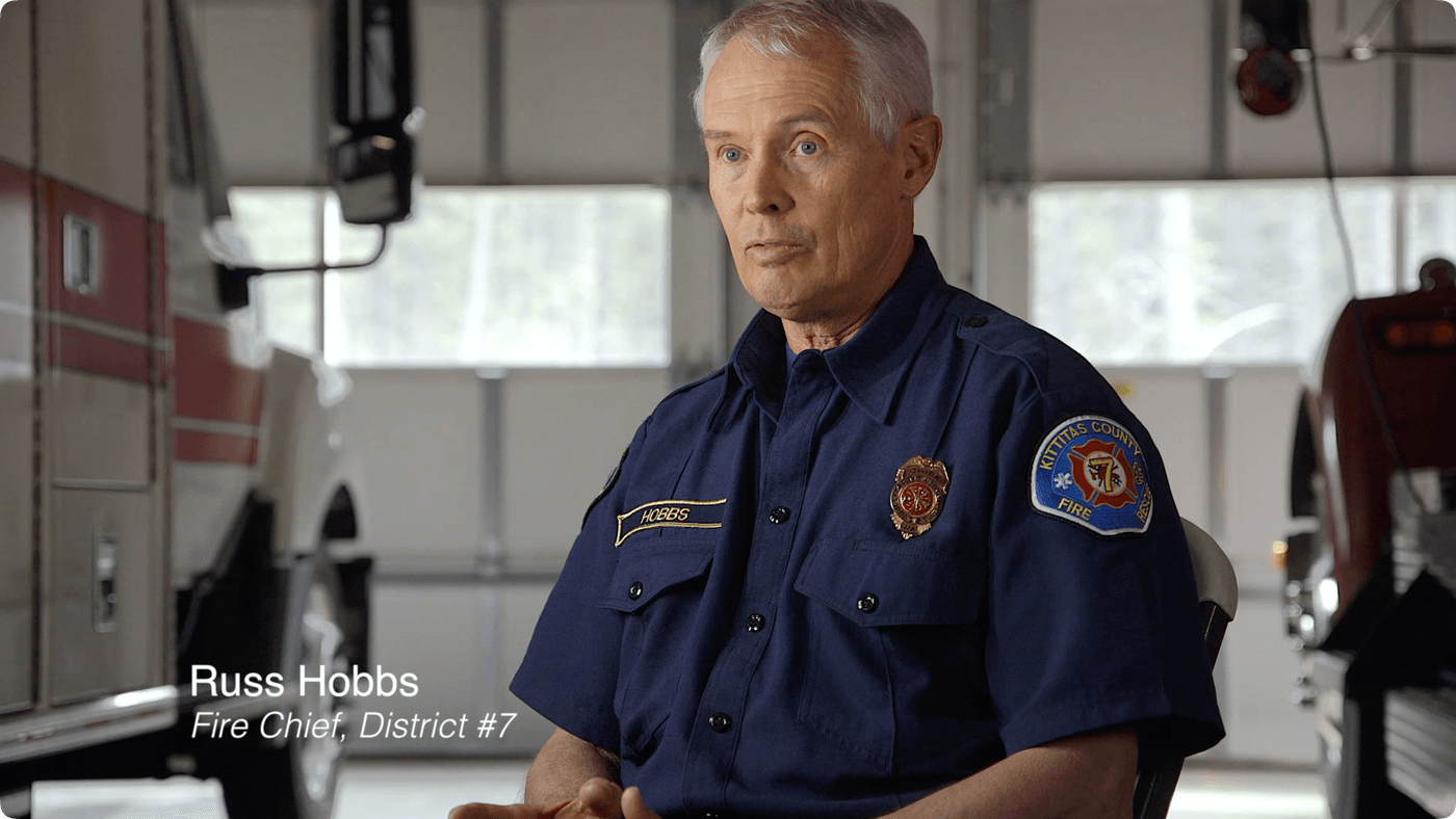 Russ Hobbs, Fire Chief for Kittitas County District #7, is centered in the frame wearing a blue uniform shirt with an embroidered patch and badge while sitting in a fire station with a fire truck in the background.