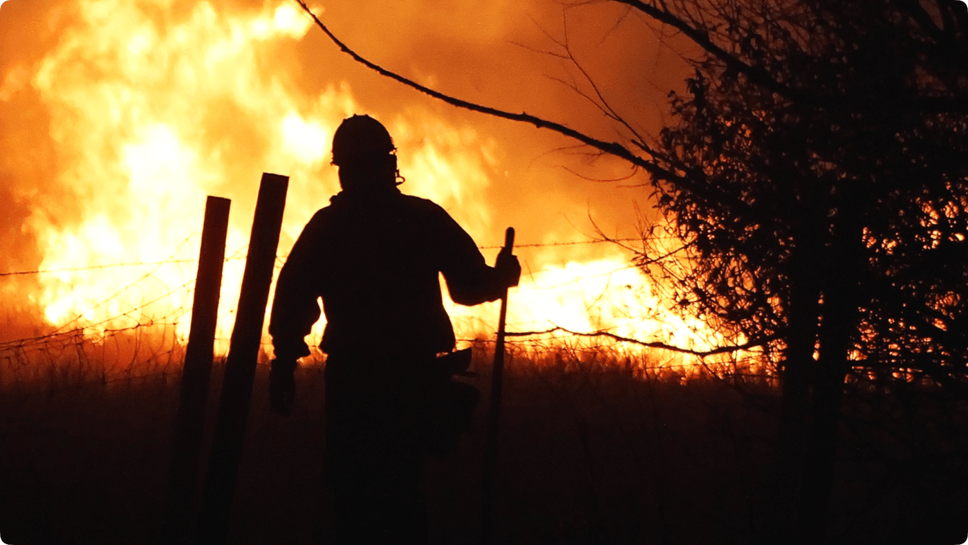 A silhouetted firefighter stands in front of a blazing wildfire at night, holding a tool near a barbed-wire fence.