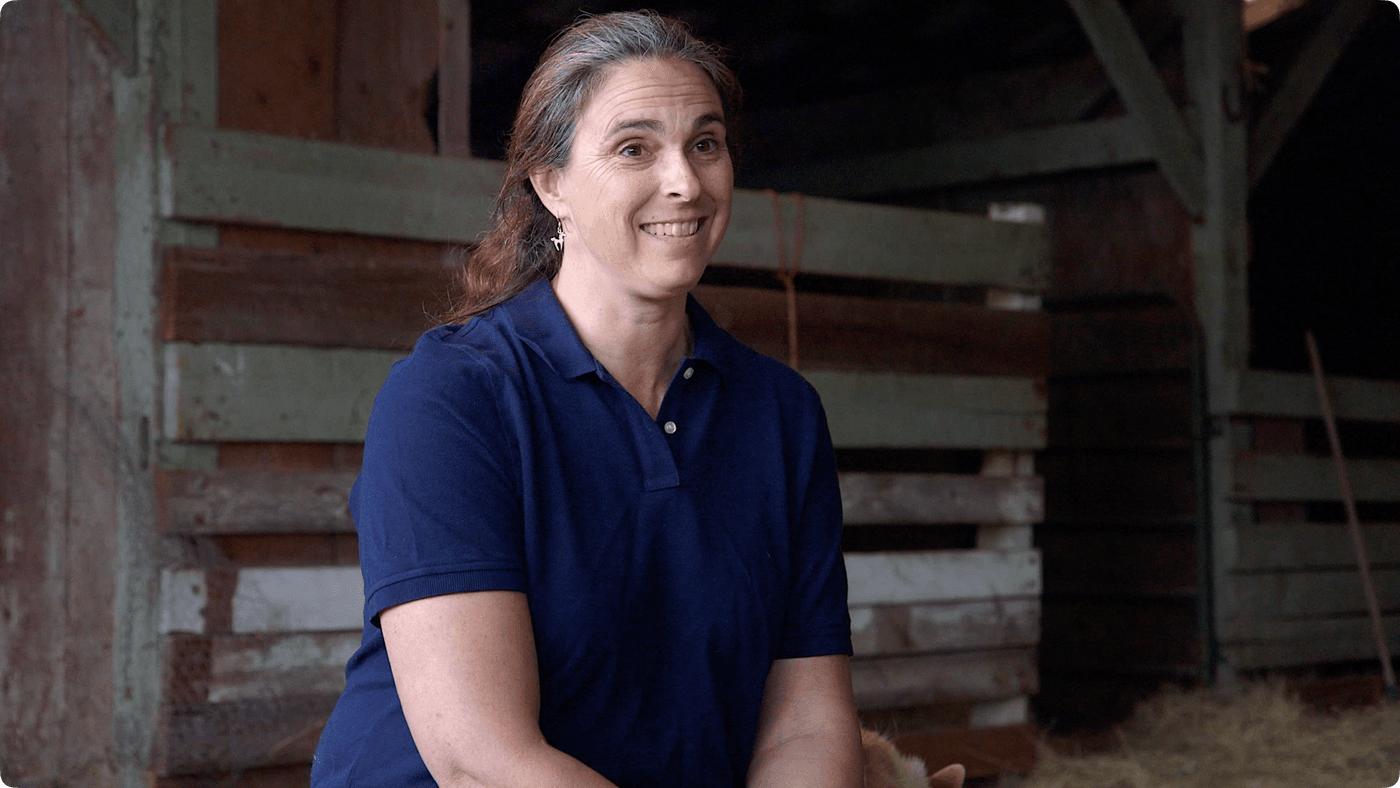 A woman with long, partially grey hair and a navy blue polo shirt smiles while sitting inside a rustic, wooden barn.