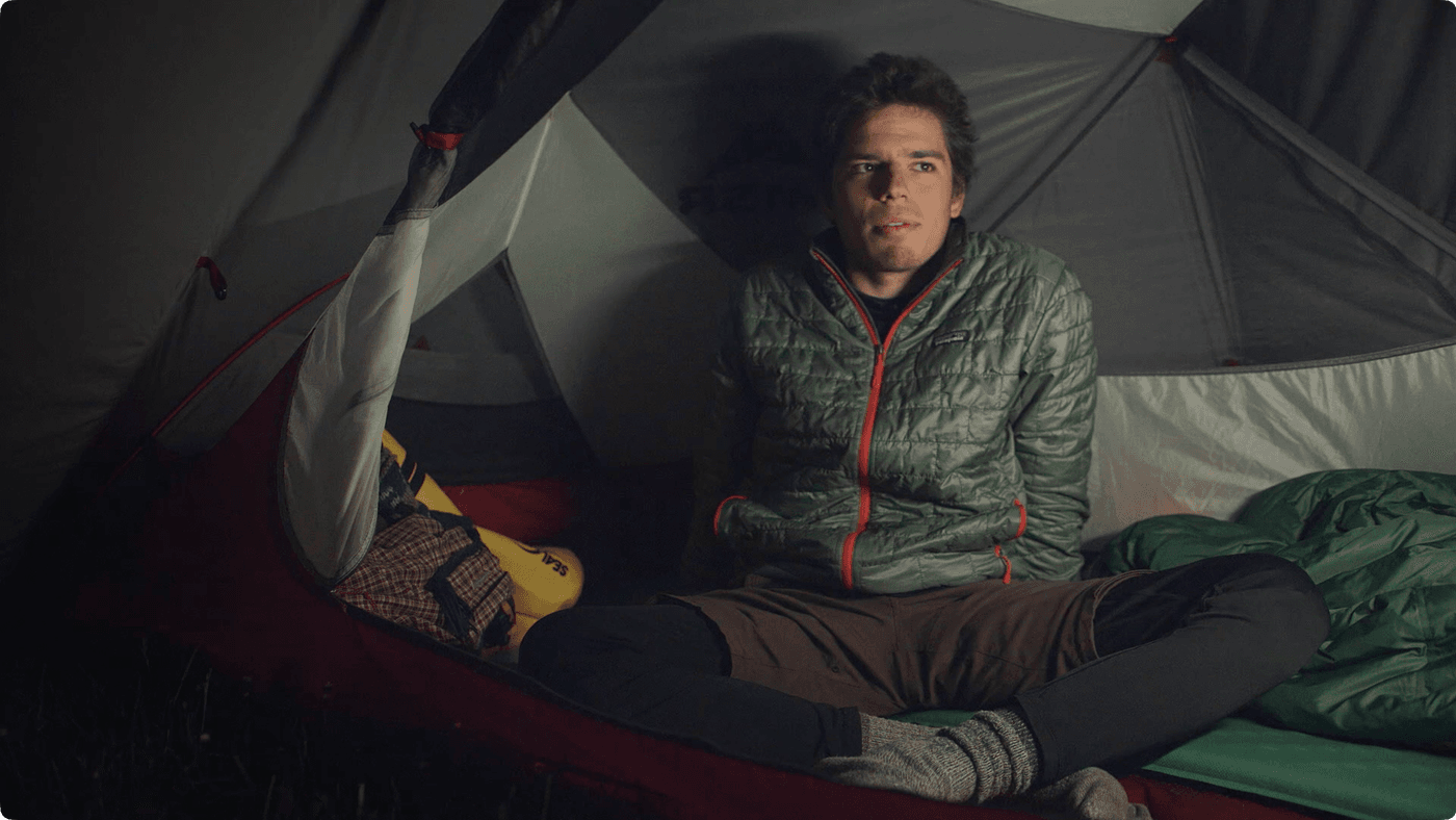 A young man wearing a green quilted jacket sits inside a camping tent at night, looking off to the side.