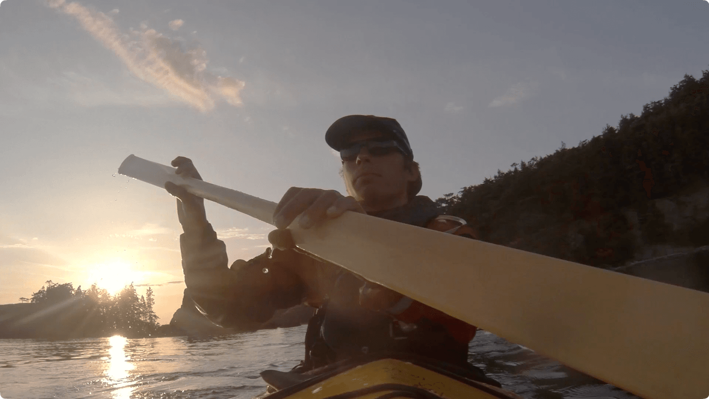 A person wearing a cap, sunglasses, and a life vest paddles a kayak across a calm body of water at sunset, with a sunlit shoreline visible in the background.