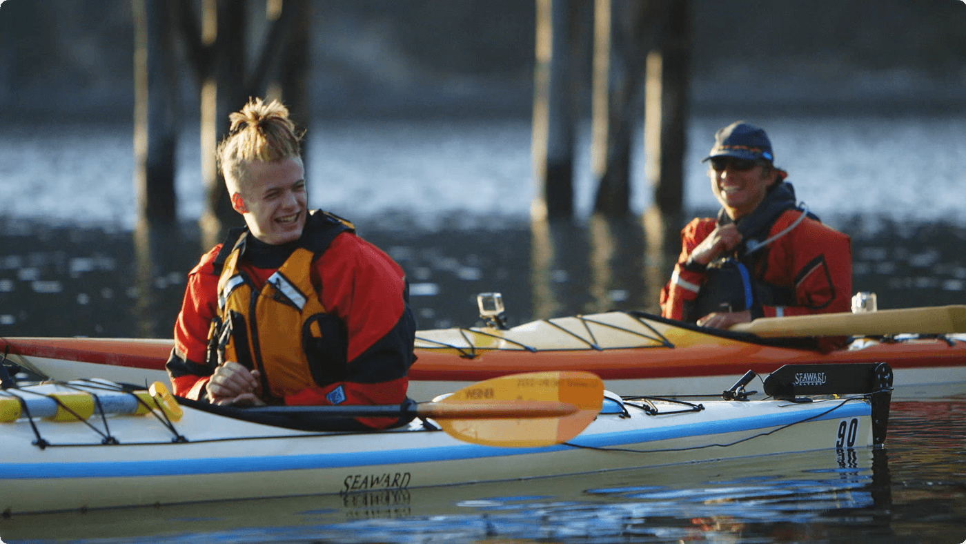 Two people in bright kayaking gear paddle tandem sea kayaks near wooden pilings in the water, both smiling and looking toward each other.