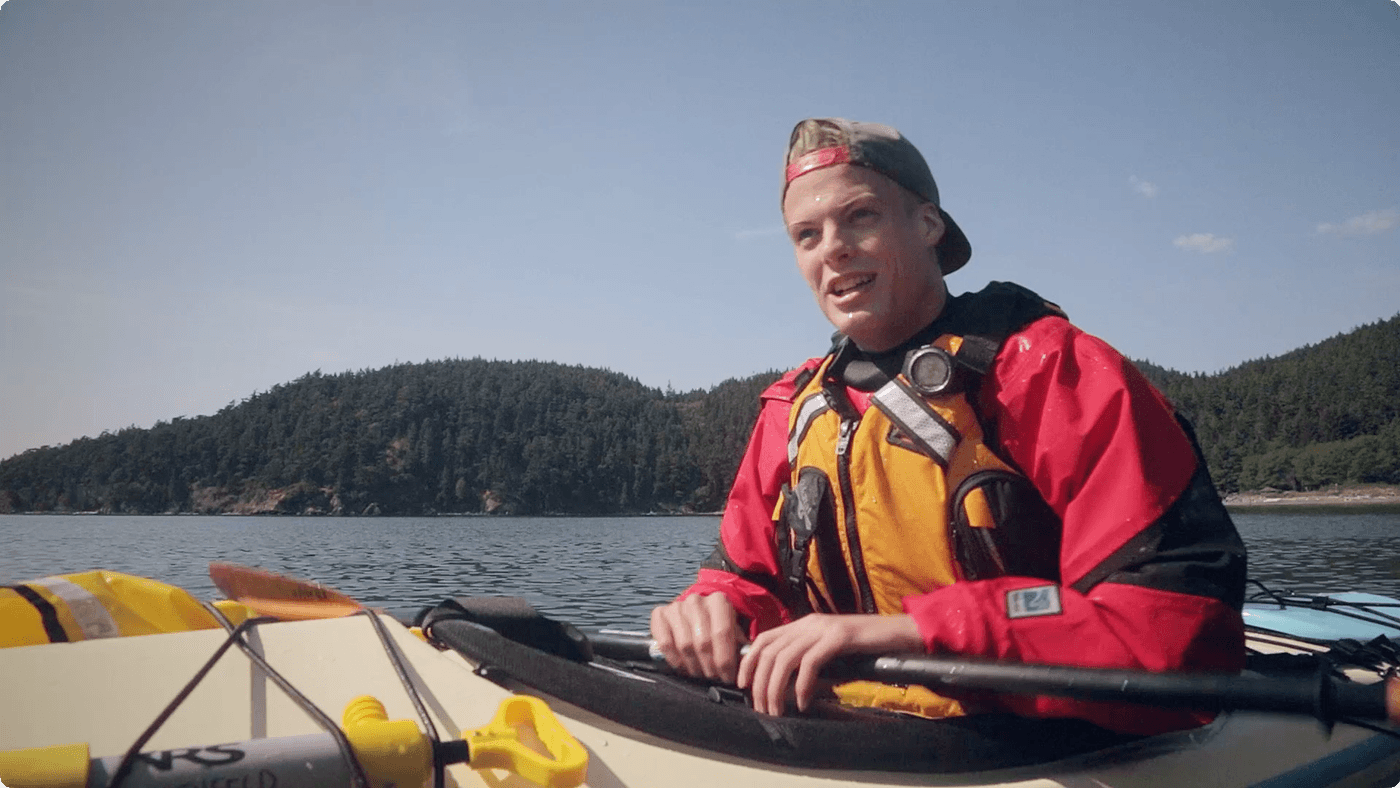 A young person wearing a red jacket, a life vest, and a backwards baseball cap paddles a kayak on a calm body of water with a forested coastline in the background.