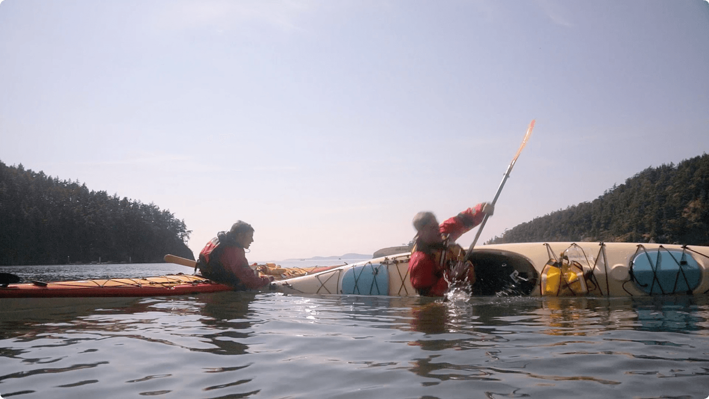 Two kayakers in red jackets practice a rescue maneuver in a calm, clear bay surrounded by forested islands, with one person in the water bracing their paddle against the other kayak.