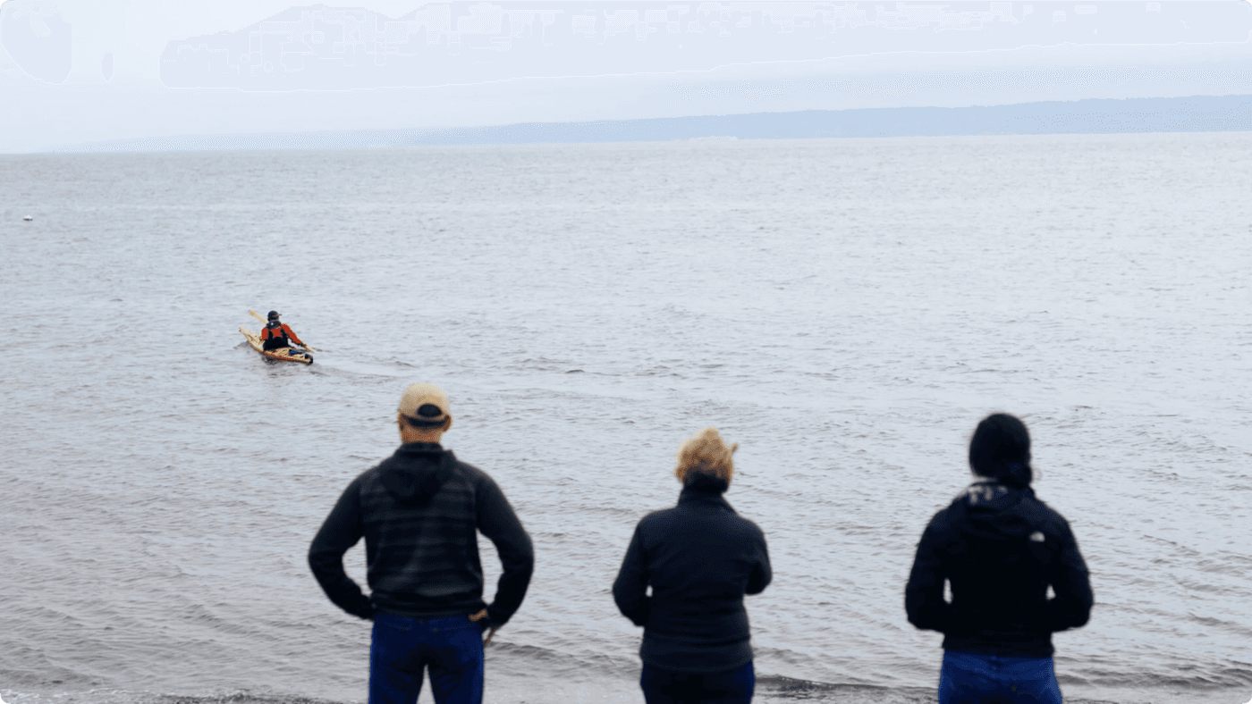 Three people stand on a shoreline with their backs to the camera, watching a person paddling a kayak across a calm, grey bay.