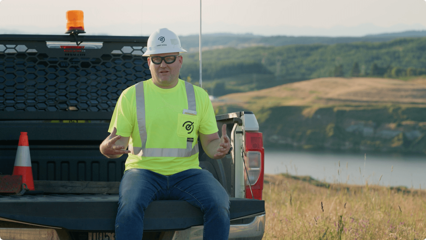 A man in a hard hat and high-visibility work shirt sits on the lowered tailgate of a pickup truck parked in a grassy landscape overlooking a river.