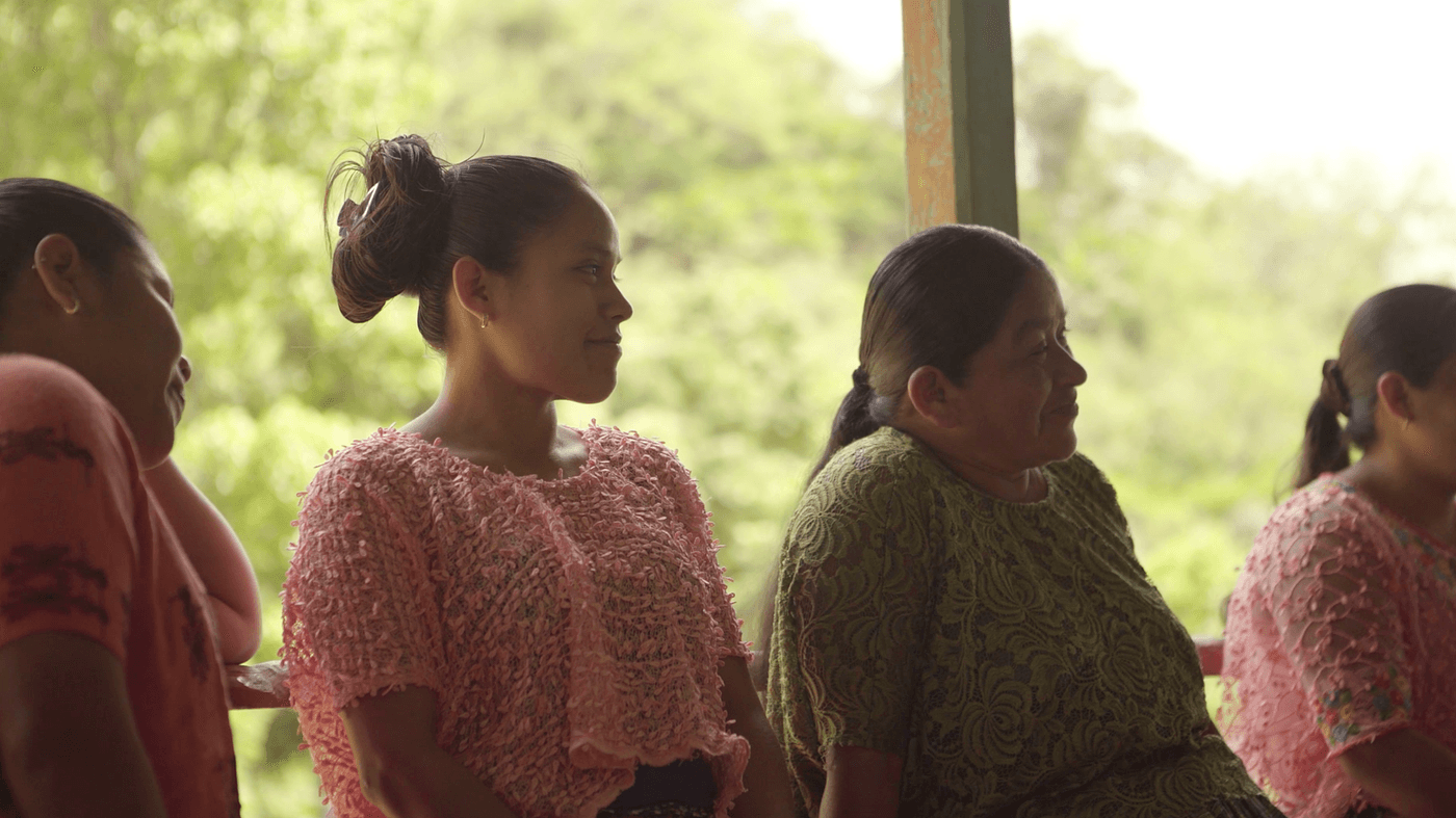 A row of four women of indigenous appearance sit together indoors, with the two in the middle wearing matching pink lace tops and the others in orange and green shirts, against a soft-focus background of lush greenery.