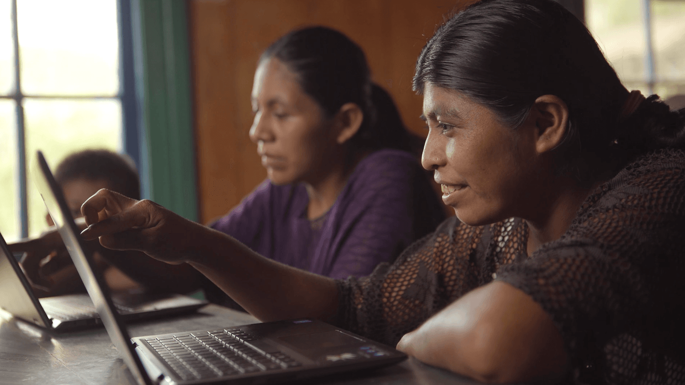 A woman with dark hair in a brown crocheted top leans over a laptop, smiling while pointing at the screen, with another woman and child working on laptops in the background of a brightly lit room.