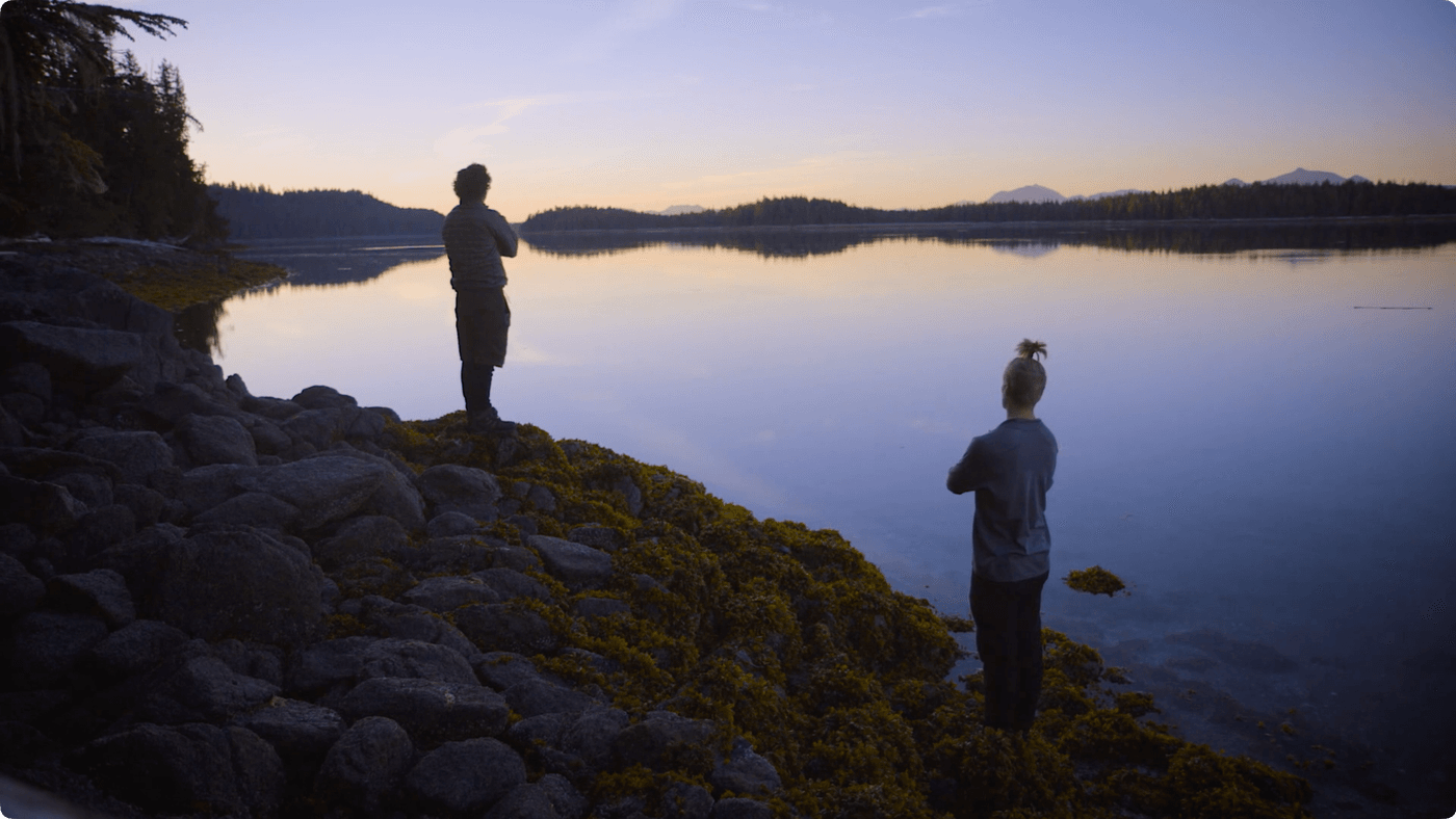Two people stand on a mossy, rocky shoreline at dusk, gazing out over a calm, reflective body of water toward distant, silhouetted mountains.