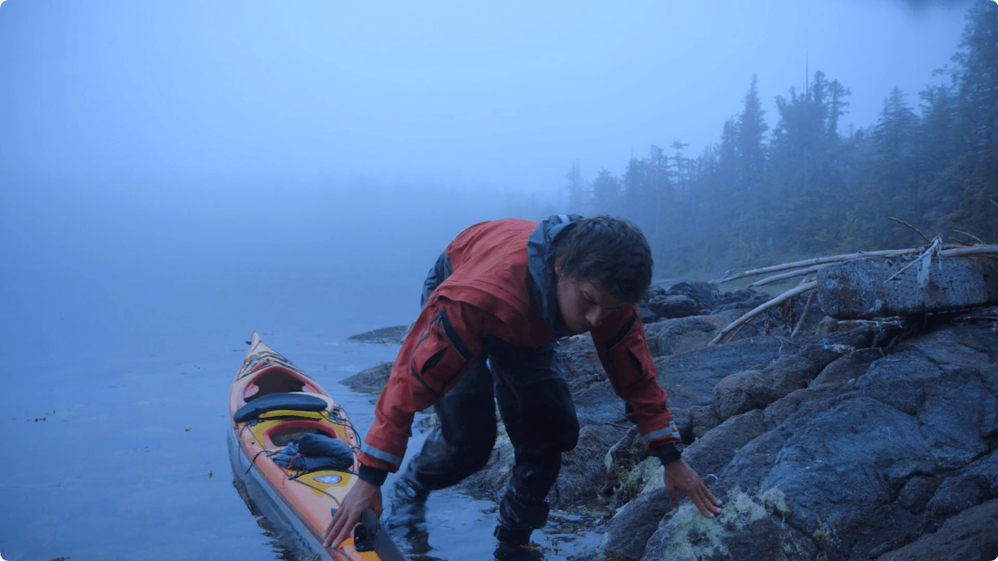 A person in a red dry suit exits an orange and yellow sea kayak onto a mossy, rocky shoreline on a foggy morning.