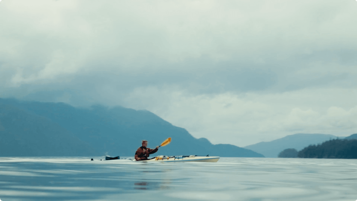 A person in a red jacket paddles a kayak across a calm, expansive body of water toward a distant mountain range under a cloudy sky.