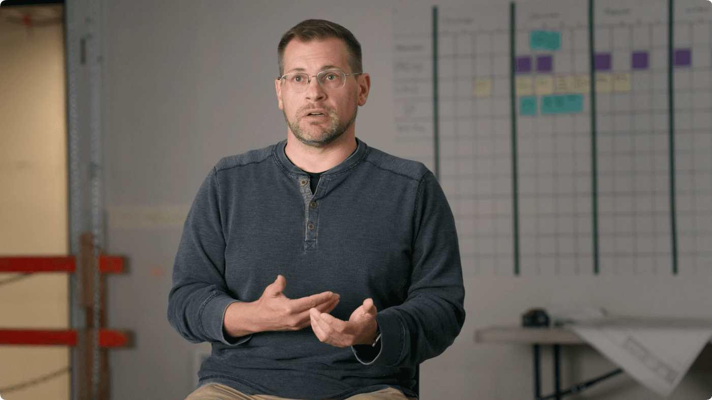 A man with glasses and a beard wears a grey long-sleeved shirt and gestures with his hands while speaking in front of a white wall with a large planning chart.