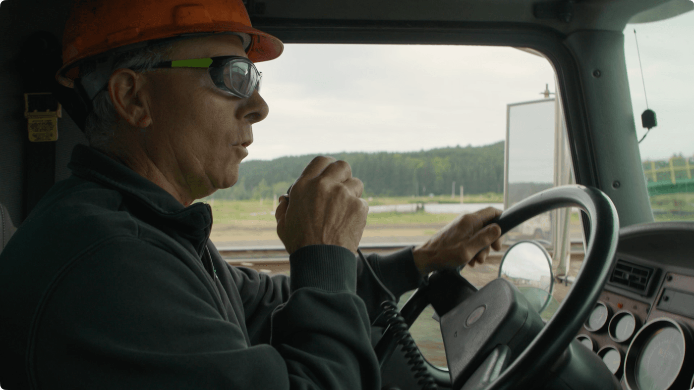 A man wearing an orange hard hat and safety glasses drives a truck while speaking into a handheld radio.