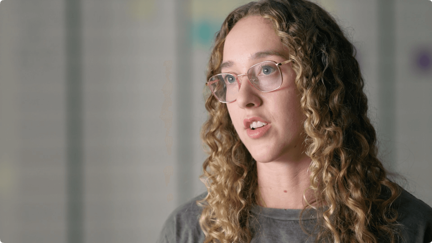 A young woman with curly blonde hair and glasses speaks while looking slightly away from the camera.
