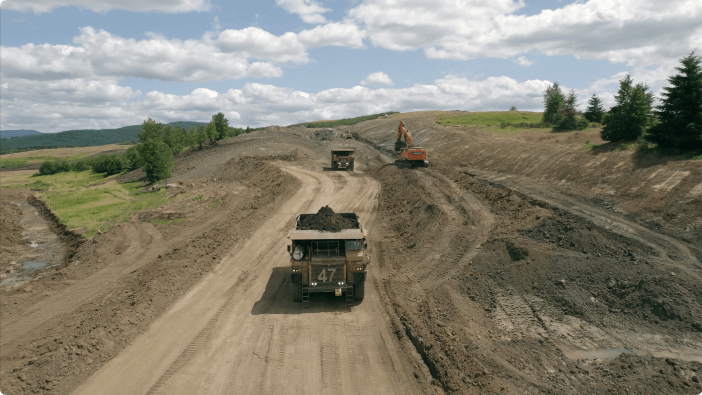 An aerial view shows two large dump trucks driving on a dirt road through a sprawling excavation site, with an orange excavator operating on a sloped hillside under a partially cloudy blue sky.