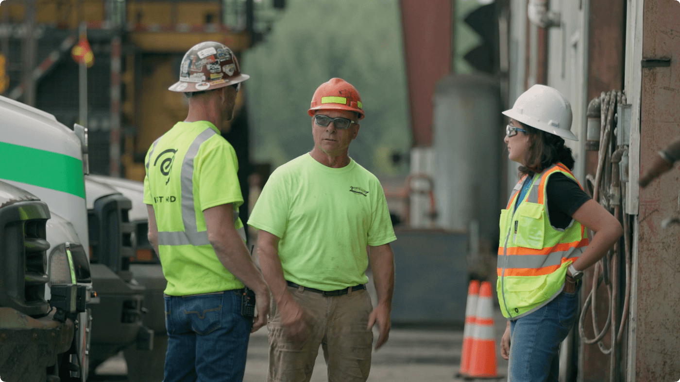 Two men in hard hats and high-visibility work shirts and a woman in a white hard hat and high-visibility vest stand outside a facility, engaged in a professional conversation.