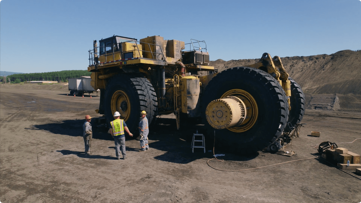 Three construction workers wearing hard hats and high-visibility gear stand on a dirt lot next to a large, yellow mining haul truck missing several wheels.