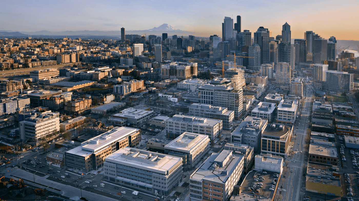 An aerial view of the Seattle skyline at sunset, featuring modern office buildings in the foreground, skyscrapers in the distance, and the snow-capped peak of Mount Rainier visible on the horizon.