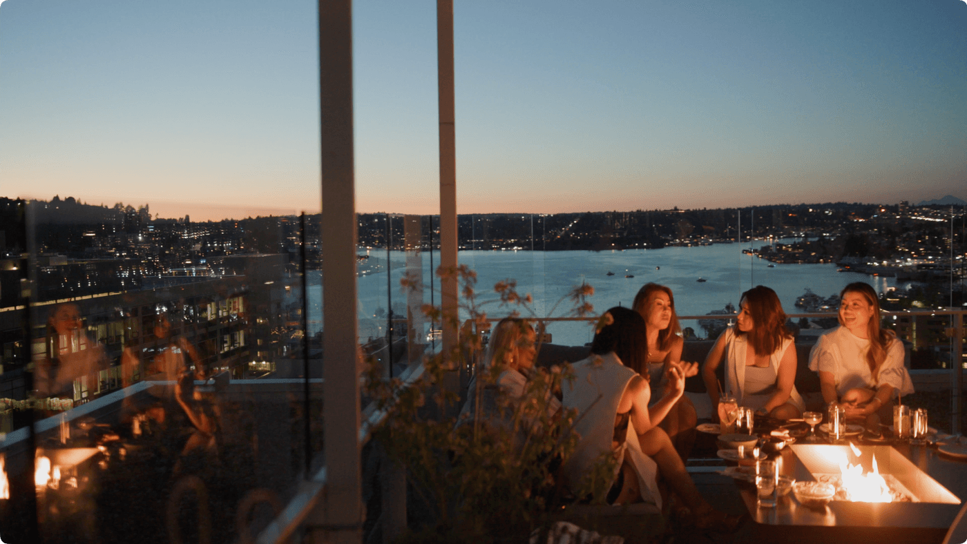 Four women sit on a rooftop terrace around a lit fire pit at twilight, overlooking a city skyline and body of water.