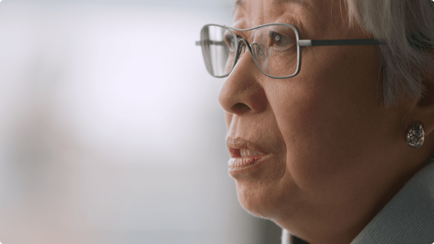 A close-up profile shot of an elderly woman with silver hair, wearing modern metal-rimmed glasses and textured silver stud earrings, looking thoughtfully toward the side.
