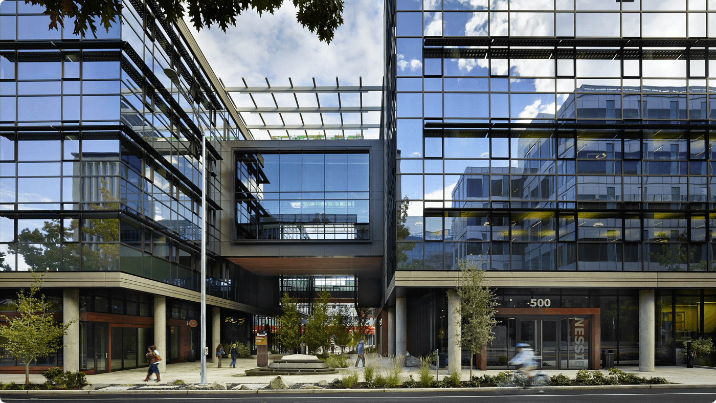 A modern office complex with glass-paneled walls features a central, elevated bridge structure connecting two buildings above a pedestrian courtyard with landscaping and a small fountain.