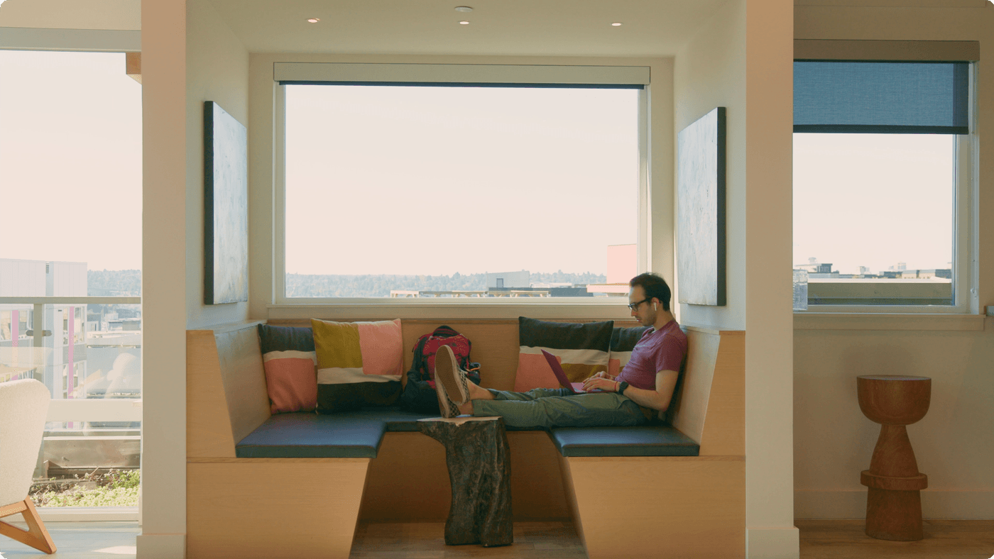 A man sits on a built-in wooden corner bench with his feet resting on a small tree-stump table while typing on a laptop, situated before a large window overlooking a city view.