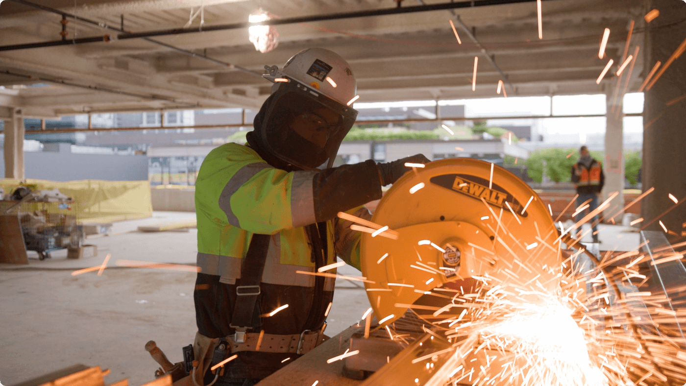 A construction worker in a high-visibility jacket and a full-face safety shield uses a yellow DeWalt chop saw to cut a metal beam, sending a spray of bright sparks into the air inside an unfinished building.