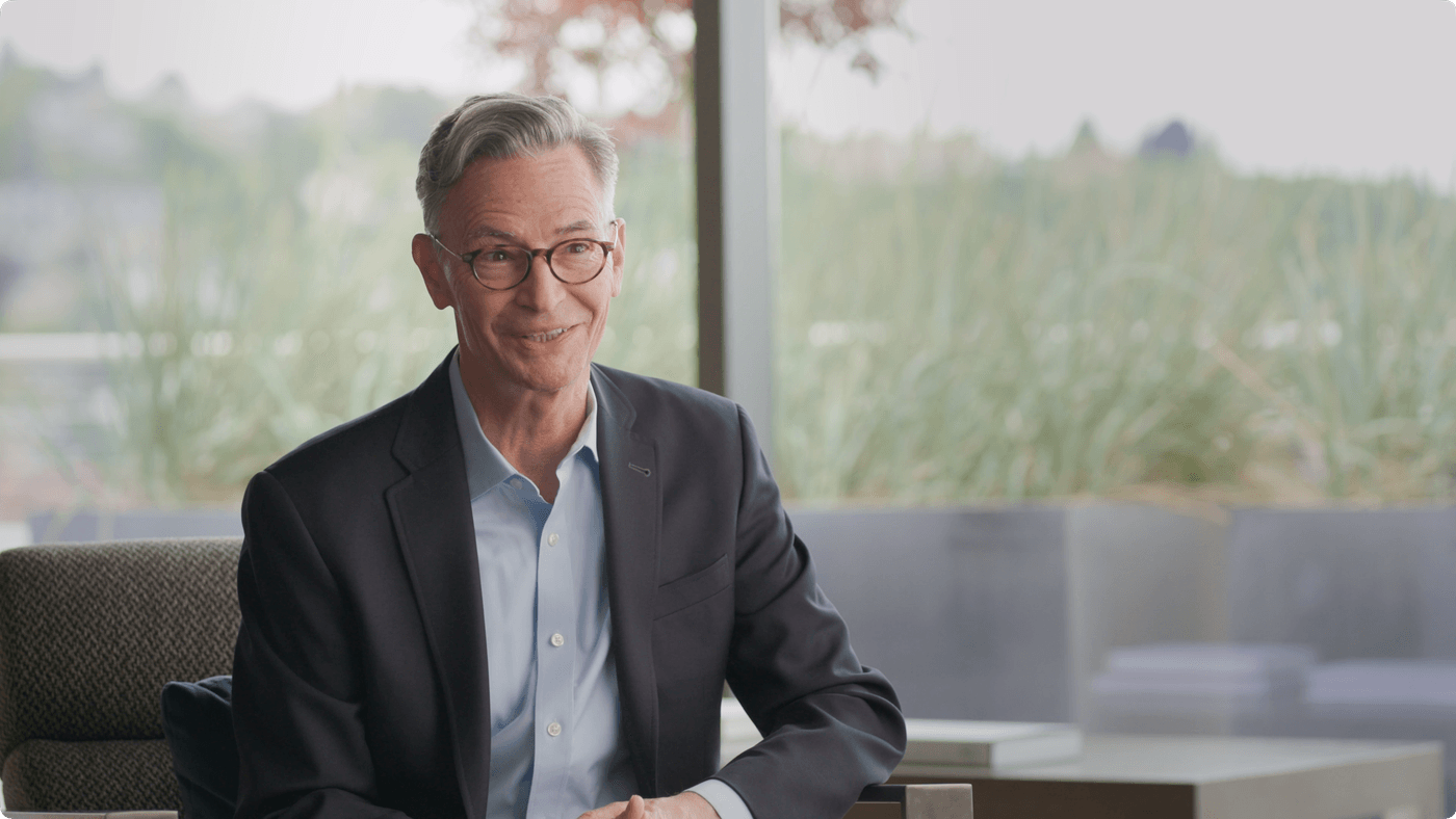 A middle-aged man with gray hair and glasses wearing a suit jacket and light blue button-down shirt sits in front of a window.