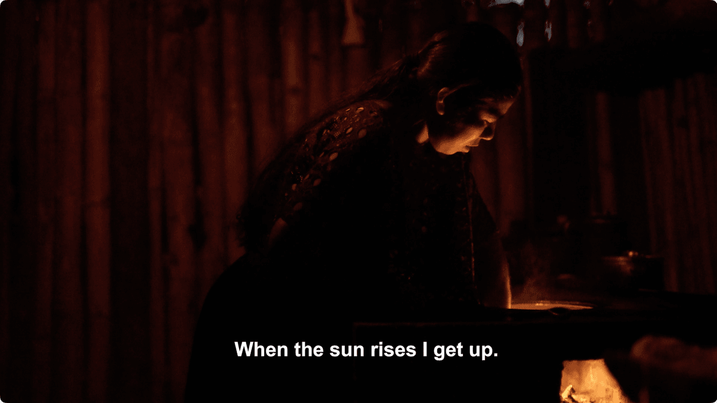 A woman with long dark hair, illuminated by a warm firelight in a dim room with bamboo walls, leans over a cooking surface while the caption reads, "When the sun rises I get up."