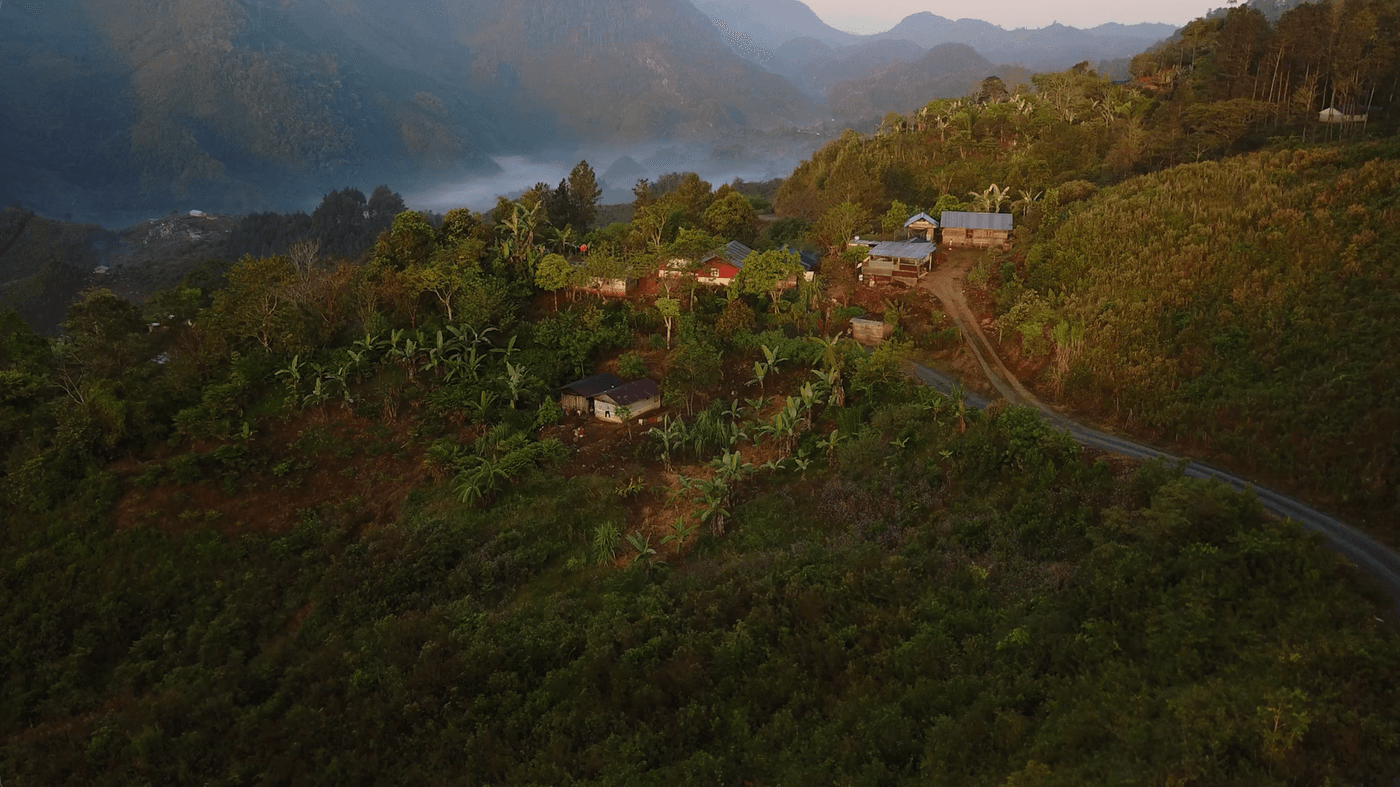 An aerial view of a small rural settlement nestled on a lush, mountainous hillside during sunrise, with a dirt path winding through the trees and mist filling the valley below.