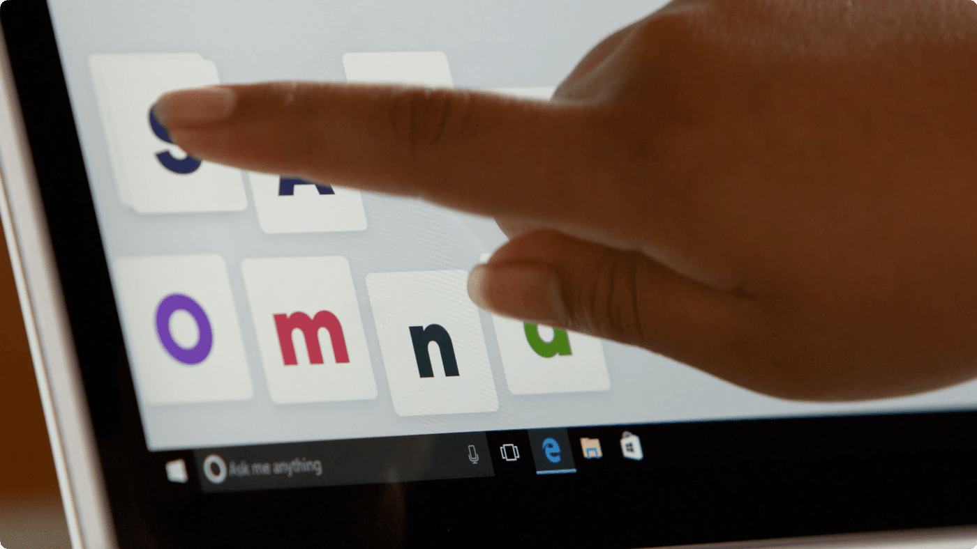 A close-up view shows a person's finger touching a touchscreen tablet displaying colorful, square tiles featuring the letters S, A, O, m, n, and d, over a Windows desktop interface.