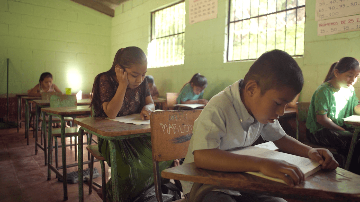 A group of young students in a brightly lit classroom are seated at wooden desks, focused on reading and studying from books and papers.