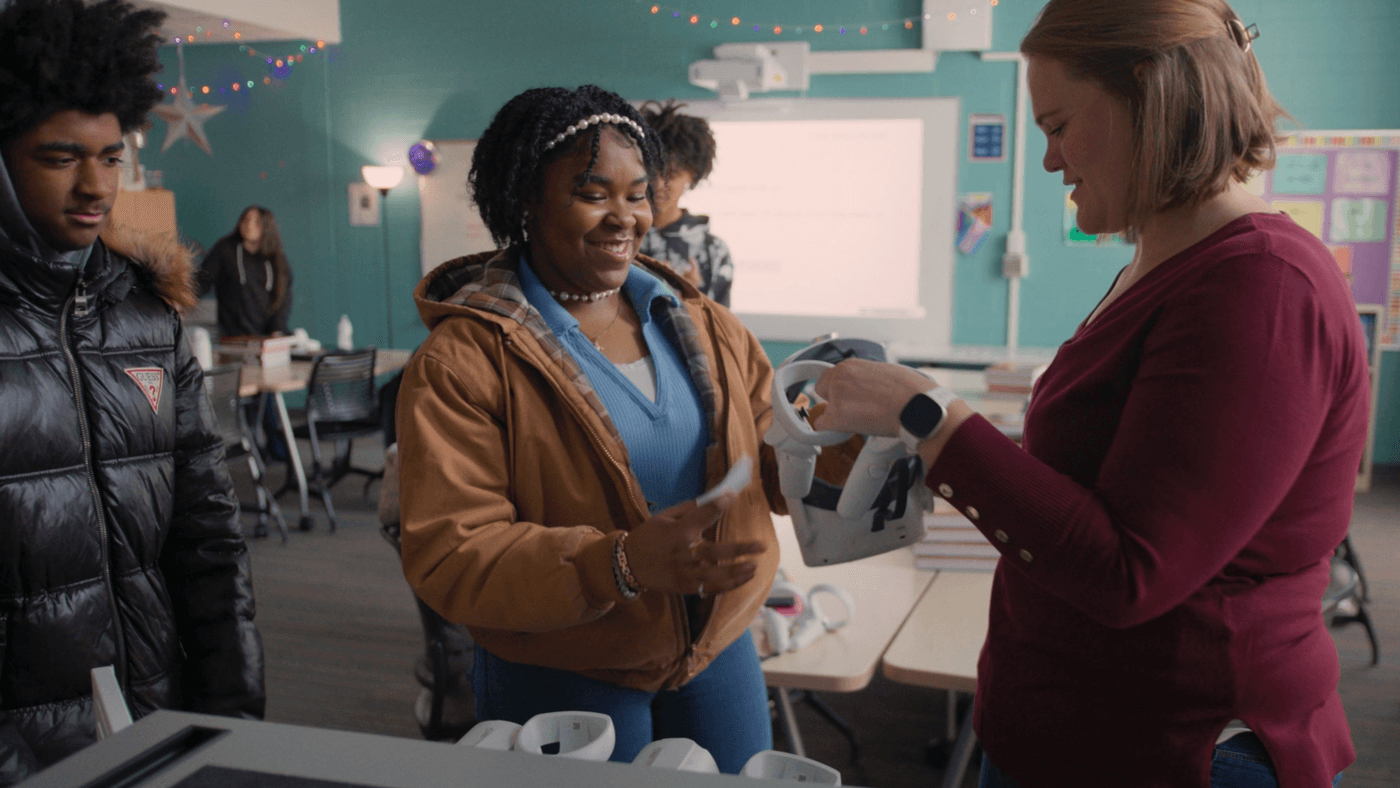 A teacher stands in a classroom handing a white virtual reality headset to a smiling female student while a male student wearing a puffy jacket watches nearby.