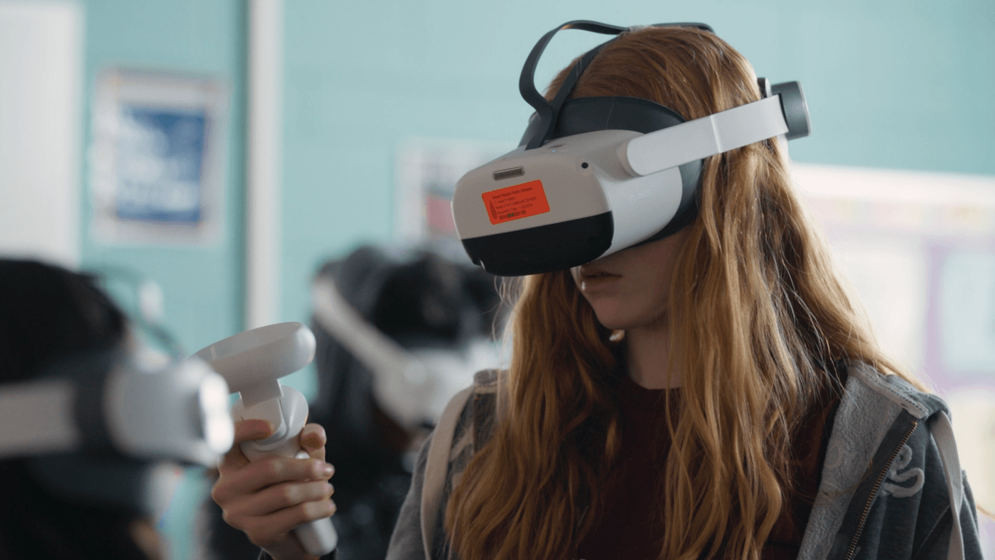 A young girl with long reddish-blonde hair wears a Pico VR headset and holds a controller while participating in a virtual reality activity in a classroom setting.
