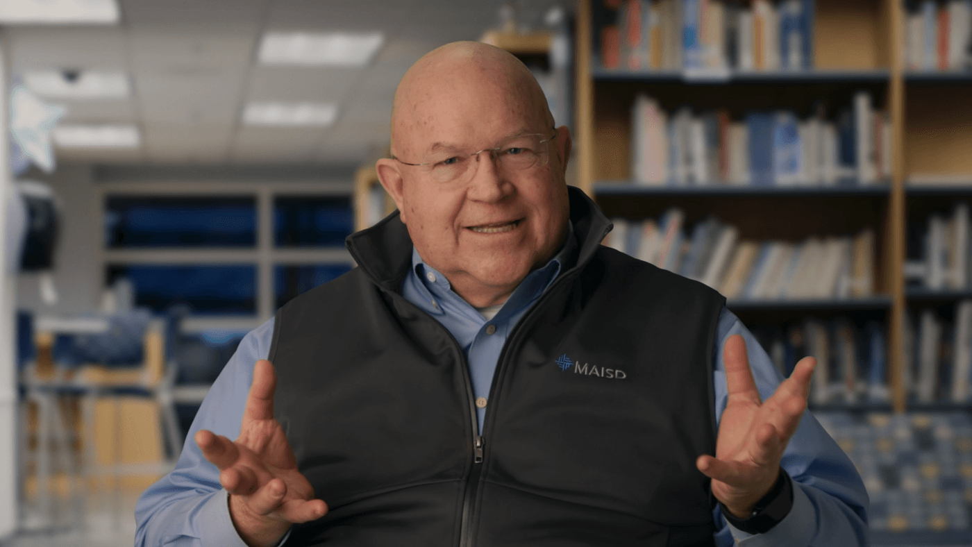 A bald man with glasses, wearing a blue button-down shirt and a dark vest with a "MAISD" logo, smiles while gesturing with both hands in front of a blurred bookshelf.