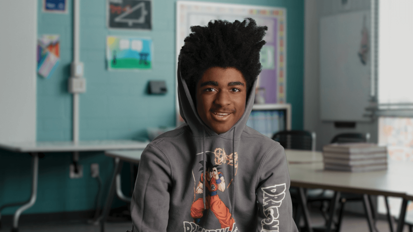A young man with voluminous, dark curly hair pulled up wears a grey Dragon Ball Z hoodie while sitting in a classroom.